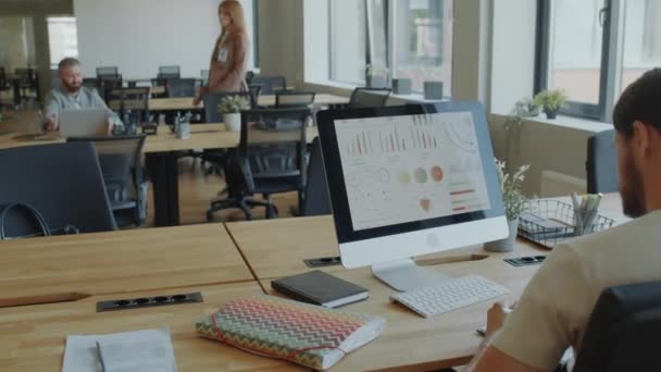 コンピュータディスプレイ上のビジネスグラフを調べ オープンスペースオフィスの仕事でノートを書き留める男の背後からの眺め — ストック動画