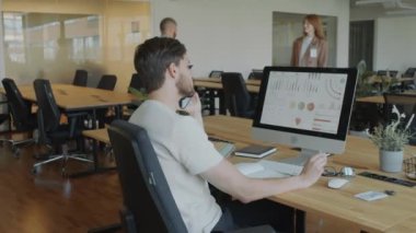 Erkek ofis çalışanı masada oturuyor, bilgisayar ekranındaki mali raporlara bakıyor, notlar yazıyor ve cep telefonuyla konuşuyor.