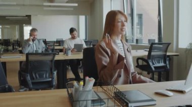 Modern açık uzay ofisindeki masada oturan ve e-toplantı sırasında bilgisayardan konuşan genç bir iş kadını.