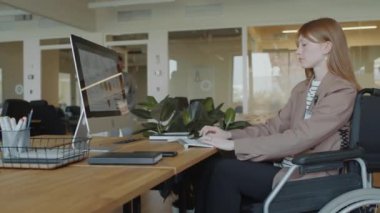 Tekerlekli sandalyedeki genç iş kadını modern açık uzay ofisindeki masaüstü bilgisayarında çalışıyor, arka planda yürüyen iş arkadaşları