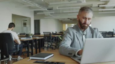 Sakallı iş adamı, açık uzay ofisinde dizüstü bilgisayarda çalışıyor.