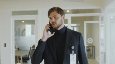 Genç işadamı modern ofiste koridorda yürüyor ve iş günü cep telefonuyla konuşuyor.