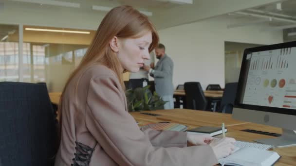 女商人在开放办公室的办公桌前工作时 分析计算机上的财务数据 并在抄本上记笔记 — 图库视频影像