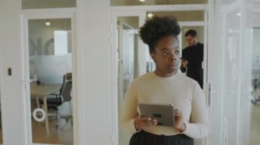 Genç Afrikalı Amerikalı iş kadını ofis koridorunda yürüyor, dijital tablet kullanıyor ve iş arkadaşları ile selamlaşıyor.