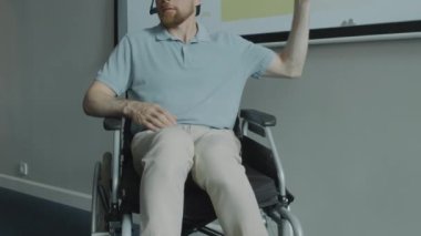Tekerlekli sandalyede oturan, sunum slaytını gösteren ve kurumsal seminer sırasında izleyicilere bilgi veren mikrofon kulaklıklı bir iş adamı görüntüsü.