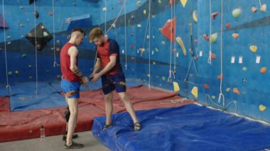 Erkek eğitmen, protetik bacaklı erkek sporcuya tırmanma antrenmanında ip takmayı öğretiyor.