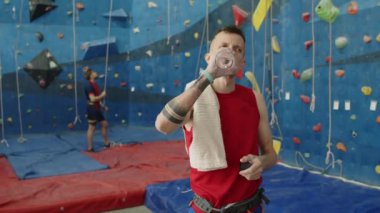 Spor kıyafetli genç adam şişeden su içiyor ve kapalı spor salonuna tırmandıktan sonra terini havluyla siliyor.