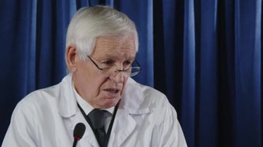 Beyaz önlüklü kıdemli doktor mavi perdeye karşı duruyor ve sağlık konferansı sırasında sahnede mikrofonla konuşuyor.