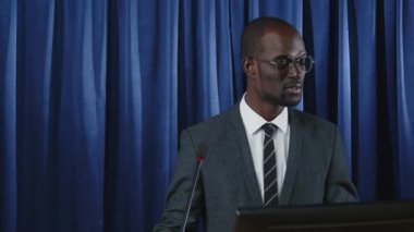 Resmi kıyafetli genç Afrikalı Amerikalı lider sahnede mavi perdeye karşı duruyor ve basın toplantısı sırasında basın açıklaması yapıyor.