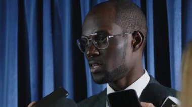 Resmi takım elbiseli genç Afrikalı Amerikalı hükümet yetkilisi konferans salonunda duruyor ve gazetecilere röportaj veriyor.