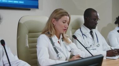 Kafkasyalı kadın doktor, çok ırklı meslektaşlarıyla masaya oturmuş mikrofonla konuşuyor ve küresel tıp konferansında açıklama yapıyor.