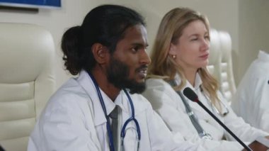 Beyaz önlüklü Hintli tıp uzmanı beyaz önlüklü kadın meslektaşıyla konferans masasında oturuyor ve halk sağlığı etkinliği sırasında mikrofonla konuşuyor.
