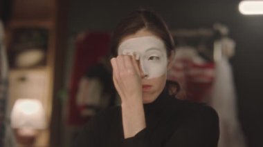 Genç bayan pandomim sanatçısı soyunma odasında oturuyor ve tiyatro gösterisine hazırlanırken yüz derisine süngerle beyaz boya sürüyor.