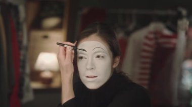 Tiyatro soyunma odasında pandomimci makyajı yaparken beyaz yüze kaşlar çizen genç bir kadın sanatçı.