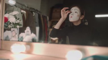 Sahne makyajlı kadın pandomimci soyunma odasındaki parlak aynaya sahte kirpik yapıştırıyor.