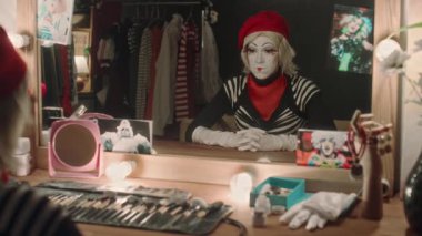 Sahne makyajlı bir aktris soyunma odasında makyaj masasında oturuyor, aynaya bakıyor ve pandomim gösterisi yapıyor.