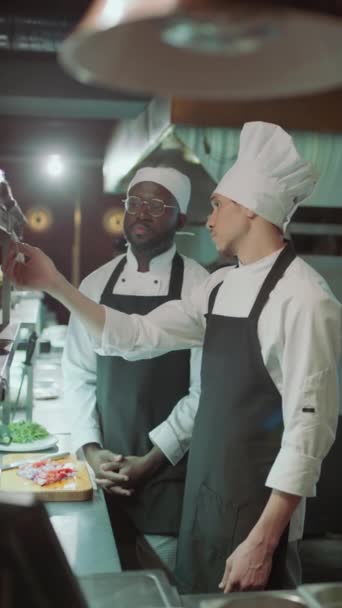 アフリカ系アメリカ人と白人男性シェフがオーダーチケットを議論し レストランのキッチンで料理をする — ストック動画