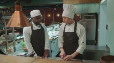 Baş aşçı malzemelerini kesiyor ve Afrika kökenli Amerikalı çırağına yemek tarifini açıklarken ona restoran mutfağında nasıl çalışacağını öğretiyor.
