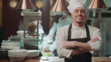 Önlüklü ve şapkalı neşeli erkek aşçının portresi kollarını kavuşturup iş günü restoranda kameraya gülümsüyor.