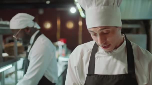 エプロンでコーカサス人の男性シェフと料理の料理を帽子で撮影し レストランのキッチンで黒い同僚と話す — ストック動画