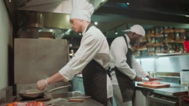 Önlüklü ve şapkalı usta aşçı tavaya alkol dolduruyor ve restoran mutfağında alevli yiyecekler pişiriyor.