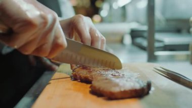 Lokanta mutfağında ahşap tahtada bıçakla kavrulmuş et bifteği kesen eldivenli erkek şefin yakın çekimleri.