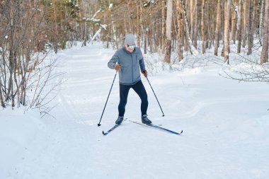 Kış günü güneş gözlüklü yaşlı beyaz adamın orman yolunda kayak yaparken çekilmiş yatay geniş bir görüntüsü.