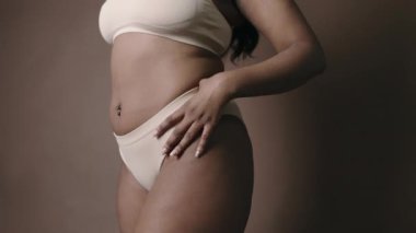 Orta bölüm stüdyo çekimlerinde tanınmayan kıvrımlı vücudu olan, minimalist iç çamaşırı giyen siyah kadın model kahve rengi arka planına karşı kameraya poz veriyor.