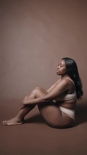 スタジオに座っている下着の若い夢のような黒人女性の垂直撮影 膝を抱き カメラのためにポーズ — ストック動画