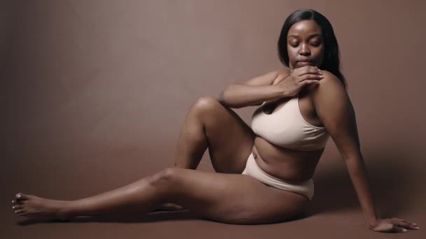 スタジオの床に座っているヌードの下着にカービーボディを着た若い女性のアフリカ系アメリカ人モデルは 彼女の肌に愛情を持って触れ カメラのためにポーズします 身体のポジティブさ 自己愛の概念 — ストック動画