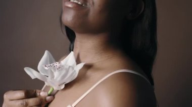 Stüdyoda poz verirken beyaz orkide çiçeğiyle tenine hafifçe dokunan sütyenli Afrikalı Amerikalı kadını yakından çek.