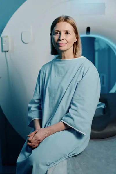 现代医院的老年白种人妇女穿着蓝色病人长袍坐在Ct扫描仪床上 垂直中弹 免版税图库图片