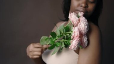 Güzel, kıvrımlı vücutlu, çıplak iç çamaşırlı, soluk pembe güllerle tenine dokunan, stüdyoda poz veren Afro-Amerikan bir kadın.