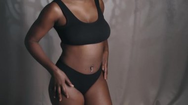 Güzel vücutlu, minimalist sütyen ve külot giymiş genç Afrikalı Amerikalı bir kadının portre fotoğrafını kaldır. Tenini okşa ve stüdyoda kameraya poz ver.