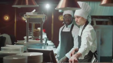 Şef restoran mutfağında yürüyor, metal raflara sipariş bileti ekliyor ve iş günü iş arkadaşlarıyla tartışıyor.