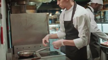 Önlüklü, şapkalı ve eldivenli bir erkek aşçı restoran mutfağında siparişi hazırlarken biftekleri tavaya koyuyor.