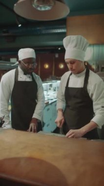 Önlüklü ve şapkalı baş aşçının dikey çekimi mutfak masası malzemelerini kesmesi ve restoranda çırakla konuşması.