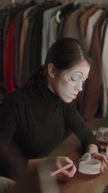 Dişi pandomim sanatçısının soyunma odasında otururken fırçayla yüzüne pudra sürerken ve gösteri için sahne makyajı yaparken dikey pozu.