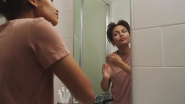 Genç, uykucu kızın banyo aynasında yansımasına bakıp sabah evde uyandıktan sonra kabarık suratını ovaladığı zoom çekimi.
