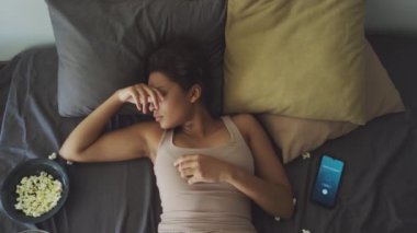 Dağınık patlamış mısırla uyanan, telefondaki alarmı kapatan ve ev partisinden sonra kendini kötü hissederken yüzünü ovuşturan genç kızın tepeden tırnağa fotoğrafı.