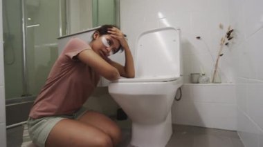 Göz altı bantlı hasta genç kız banyoda yerde oturuyor, tuvalete yaslanıyor ve gıda zehirlenmesinden acı çekerken derin nefes alıyor.