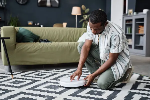 Junger Afroamerikaner Sitzt Wohnzimmer Auf Dem Boden Und Schaltet Staubsaugerroboter Stockfoto