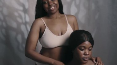 Minimalist iç çamaşırlı iki güzel siyahi kadının, stüdyoda gülümseyerek kamera önünde poz verişini aşağı indirin.