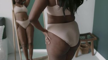 Pastel sutyen ve külot giyen siyah kadın aynaya bakıyor ve evdeki güzel gününün tadını çıkarıyor.