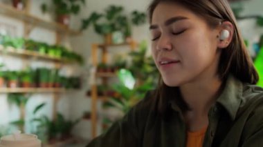 Bilgisayarında kablosuz kulaklıkları olan meşgul, genç, beyaz bir kadının telefonuyla müşterilerle elleri olmadan konuşurken görüntüsü. Ahşap raflarda yeşil saksılarla süslenmiş duvarlar.