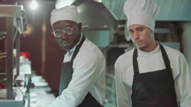 Şef restoran mutfağında yürüyor, sipariş biletini alıyor ve rafa koyuyor, sonra da erkek iş arkadaşına iş veriyor.