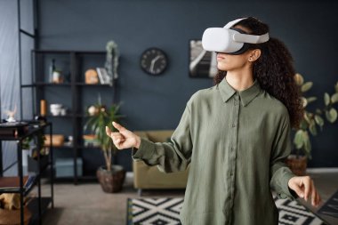 Modern melez bir kızın evde dikilip VR kulaklık taktığı orta boy bir fotoğraf.