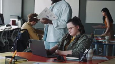Down sendromlu genç bir iş kadını çok ırklı meslektaşlarıyla birlikte açık uzay ofisinde dizüstü bilgisayarda çalışıyor.