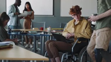 Tekerlekli sandalyedeki genç işadamı dizüstü bilgisayarda çalışıyor ve ofiste protez bacaklı bir meslektaşla belgeyi tartışıyor.