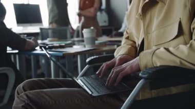 Tekerlekli sandalyede oturan ve ofiste çalışırken dizüstü bilgisayarda yazan engelli genç adamın yakın görüntüsü
